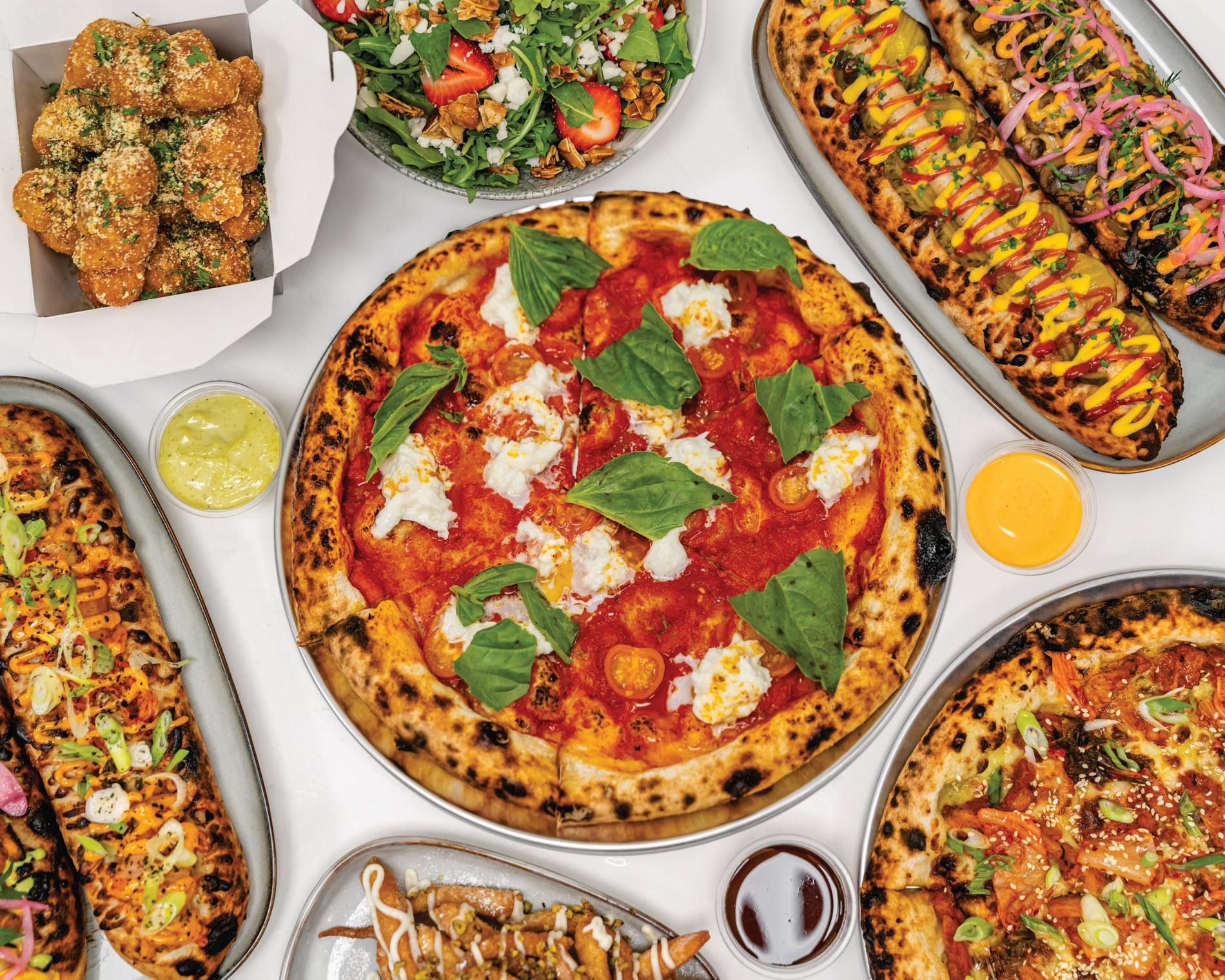 A spread of Baked Columbia pizzas including a margherita with burrata and basil, loaded flatbreads, garlic knots, salad and dips